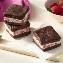 Imagem aproximada da receita de Sanduíche de Brownie com Sorvete, em quadrados marrons, recheados com um creme claro com pedaços de morango, todos sobre uma tábua clara em uma bancada decorada com mais morangos e um tecido em tom rosa