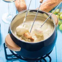 Foto da Receita de Fondue de Queijo Sem Lactose. Observa-se uma panelinha de fondue azul com três espátulas de pão dentro.