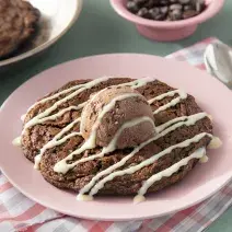 Fotografia em tons de marrom em uma bancada de madeira de cor verde. Ao centro, um pano rosa quadriculado com o cookie dentro. Ao fundo, uma frigideira contendo mais cookie e ao fundo, um pote com gotas de chocolate.