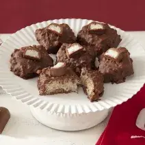 Foto da receita de cocada de prestígio. Observa-se uma boleira branca com 6 cocadas banhadas no chocolate. A da frente está cortada e é possível ver o coco de dentro