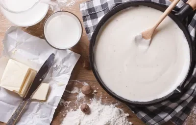 Fotografia em tons de marrom com uma panela preta ao centro. Dentro da panela existe um molho branco. Ao lado existe uma manteiga em tablete cortada em fatias.