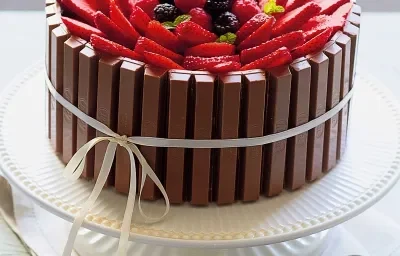 Fotografia em tons de branco de uma bancada branca com um paninho branco, um suporte para bolo branco e um bolo de Kitkat com frutas vermelhas. Ao lado um recipiente redondo branco com frutas vermelhas.