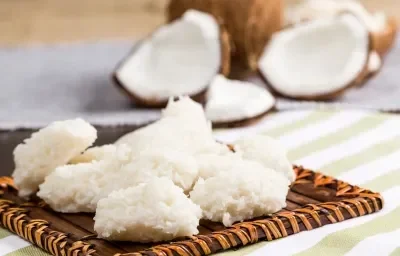 Foto da receita de Cocada Zero Lactose. Observa-se as cocadas dispostas em uma tábua e atrás é possível ver os cocos abertos.