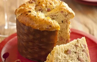 Fotografía en tonos rojos y marrones, en el centro un plato rojo con un panettone en rodajas, al lado de dos vasos con bebidas y una decoración navideña, al fondo dos platos apilados, todo sobre una rústica encimera de madera.