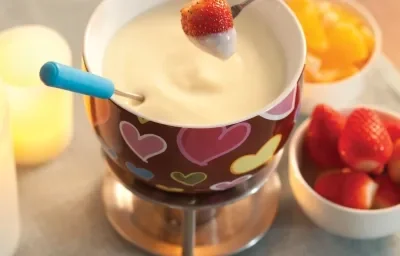 Fotografia em tons de cinza, vermelho e amarelo, com aparelho para fondue com fondue de chocolate branco com morango suspenso, no lado direito potinho com morango e outro com frutas cortadas, decoração amarela do lado esquerdo, tudo sobre toalha cinza.