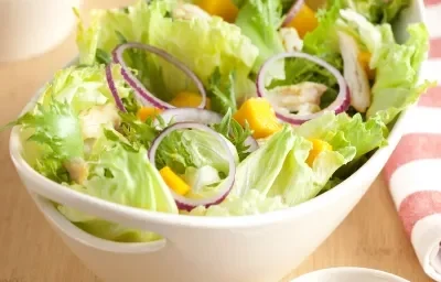 em uma mesa de madeira contém um recipiente branco que comporta a salada. Ao lado um pote com tempero e um pano listrado nas cores branco e rosa.