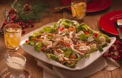 Fotografia em tons de vermelho em uma bancada de madeira escura, um pano bege, um prato branco grande com salada de alface, rúcula, lentilha e chester. Ao lado, copos, molho e enfeites e decoração natalina.