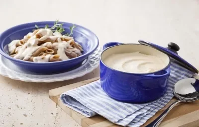 Foto de uma panela azul com molho branco dentro, em cima de uma tábua de madeira e um pano azul com branco. Do lado esquerdo, um prato de macarrão com molho branco em cima