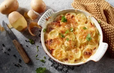 Foto da receita de Batata Gratinada Zero lactose. Observa-se um refratário redondo branco de porcelana com as batatas gratinadas dentro. Ao lado esquerdo, batatas descascadas decoram