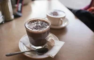 Fotografia em tons de marrom em uma bancada de madeira de cor marrom. Ao centro, um pires branco com uma xícara em cima contendo a bebida com uma colher pequena ao lado. Ao fundo, há uma xícara branca contendo a bebida.