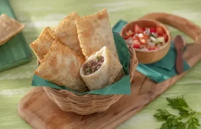 Foto da receita de Massa de Pastel Sem Glúten. Observa-se uma cestinha com 5 pastéis, sendo que um está cortado no meio mostrando o recheio de carne. Ao lado direito, uma porção de vinagrete para acompanhar.