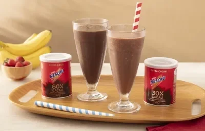 Foto em tons de vermelho da receita de shake saudável feita com Nescau Extra Cacau servida em dois copos altos de vidro sobre uma tábua de madeira com duas latas de nescau e canudos listrados. Ao fundo, bananas e morangos para decorar  sobre uma mesa bege