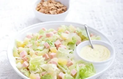 Fotografia em tons de branco e verde de uma bancada de madeira branca com paninho branco, sobre ele um recipiente redondo branco com a salada tropical e com o molho de iogurte. Ao fundo um recipiente redondo branco com o Nesfit Cereal.