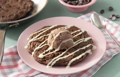 Fotografia em tons de marrom em uma bancada de madeira de cor verde. Ao centro, um pano rosa quadriculado com o cookie dentro. Ao fundo, uma frigideira contendo mais cookie e ao fundo, um pote com gotas de chocolate.