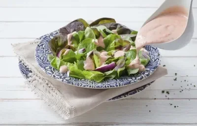 Fotografia em tons de azul, rosa e verde de uma bancada de madeira branca com paninho branco e um prato azul desenhado, sobre ele uma salada de alface com o molho rose e um recipiente despejando o molho sobre a salada.