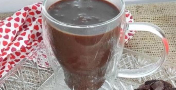 Foto da receita de Chocolate Quente. Observa-se uma xícara branca transparente com a bebida dentro, sobre um pires transparente de vidro.