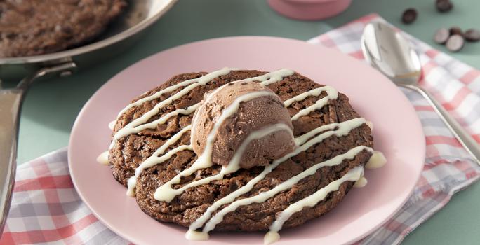 Fotografia em tons de marrom em uma bancada de madeira de cor verde. Ao centro, um pano rosa quadriculado com o cookie dentro. Ao fundo, uma frigideira contendo mais cookie e ao fundo, um pote com gotas de chocolate.