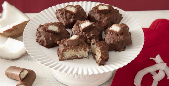 Foto da receita de cocada de prestígio. Observa-se uma boleira branca com 6 cocadas banhadas no chocolate. A da frente está cortada e é possível ver o coco de dentro