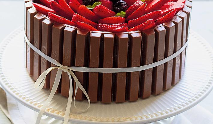 Fotografia em tons de branco de uma bancada branca com um paninho branco, um suporte para bolo branco e um bolo de Kitkat com frutas vermelhas. Ao lado um recipiente redondo branco com frutas vermelhas.