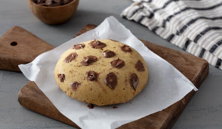 Fotografia em tons de marrom em uma bancada de madeira de cor cinza. Ao centro, uma tábua de madeira com um pedaço de papel manteiga em cima contendo o cookie. A fundo, há um pote com pedaços de chocolate e um pano branco listrado.