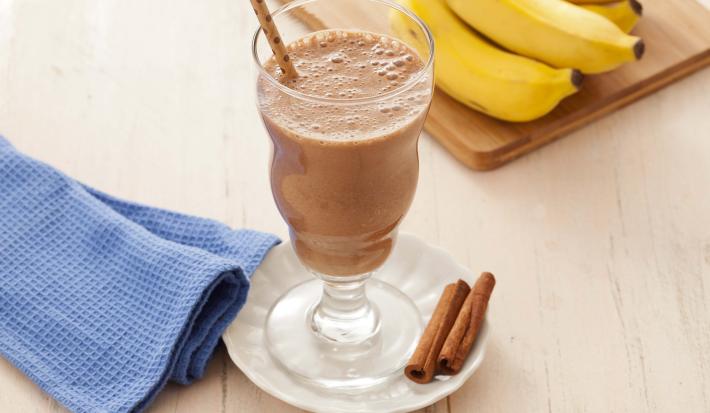 Fotografía en tonos marrones en un banco de madera clara con un paño azul al lado y en el centro un pequeño plato blanco con una taza de vidrio y el batido de cacao y plátano adentro. Al fondo, un racimo de plátanos apoyado en una tabla de madera.