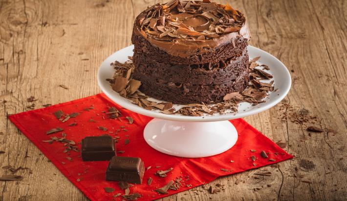 Foto da receita de Bolo Brownie com Brigadeiro. O bolo em tom marrom escuro está em cima de um prato branco mais alto, sobre uma bancada de madeira decorada com raspas de chocolate