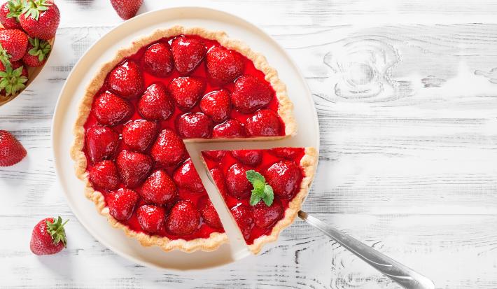 Fotografia vista de cima de um prato grande branco servindo como apoio para uma torta de morango zero lactose, com morangos por cima e uma calda brilhante. Tem um talher tirando um pedaço perfeito, e ao redor da torta tem vários morangos.