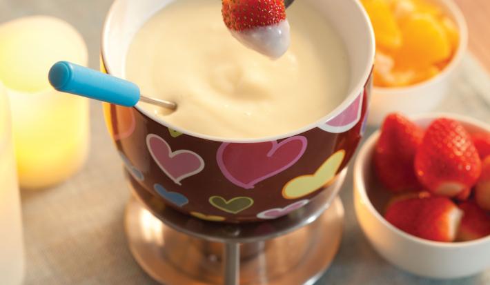 Fotografia em tons de cinza, vermelho e amarelo, com aparelho para fondue com fondue de chocolate branco com morango suspenso, no lado direito potinho com morango e outro com frutas cortadas, decoração amarela do lado esquerdo, tudo sobre toalha cinza.