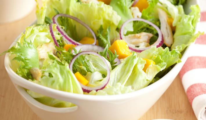 em uma mesa de madeira contém um recipiente branco que comporta a salada. Ao lado um pote com tempero e um pano listrado nas cores branco e rosa.