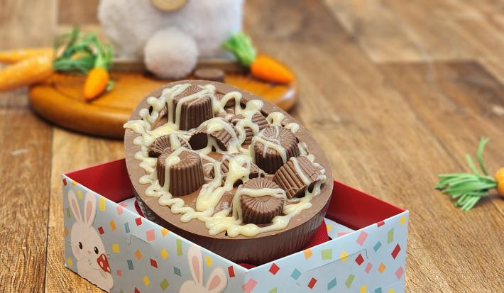 Fotografia em tons de laranja e marrom em uma bancada de madeira e uma caixa ao centro. Dentro da caixa existe um ovo de chocolate recheado com bombons e decorado com calda de chocolate branco. Ao fundo um coelho de pelúcia com cenouras.