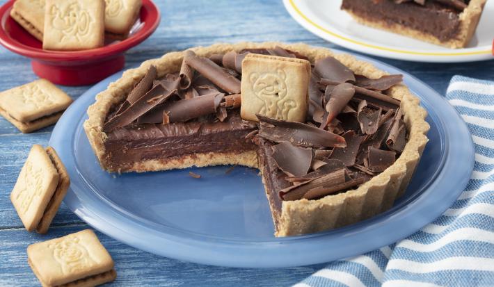 Fotografia em tons de azul em uma bancada de madeira azul, um pano azul listrado, um prato redondo raso azul com a torta de Biscoito Passatempo em cima dele, já com uma fatia cortada. Ao fundo, um prato com a fatia da torta e um potinho com os biscoitos.