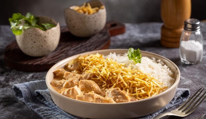 Fotografia em tons de cinza com um prato fundo ao centro. Dentro do prato existe uma receita de frango com cubos com molho de cor rosé, batata palha e arroz. Ao fundo existe um potinho com sal.