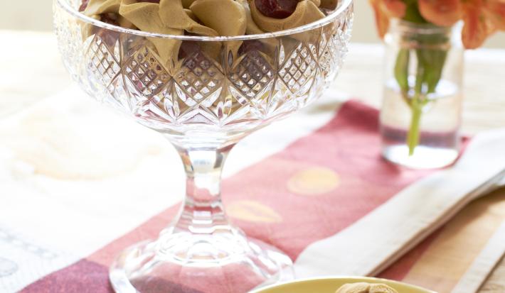 Fotografia em tons de marrom em uma bancada de madeira escura com um pano branco com detalhes vermelhos, um prato amarelo com três biscoitinhos com goiabada. E ao centro, uma bomboniere com vários biscoitinhos com goiabada.