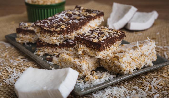 Fotografia em tons de bege em uma bancada de madeira com um jogo americano de palha, um recipiente retangular bege com algumas barrinhas de coco com chocolate em cima dele. Ao lado, pedaços de coco espalhados pela bancada.