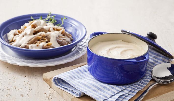 Foto de uma panela azul com molho branco dentro, em cima de uma tábua de madeira e um pano azul com branco. Do lado esquerdo, um prato de macarrão com molho branco em cima