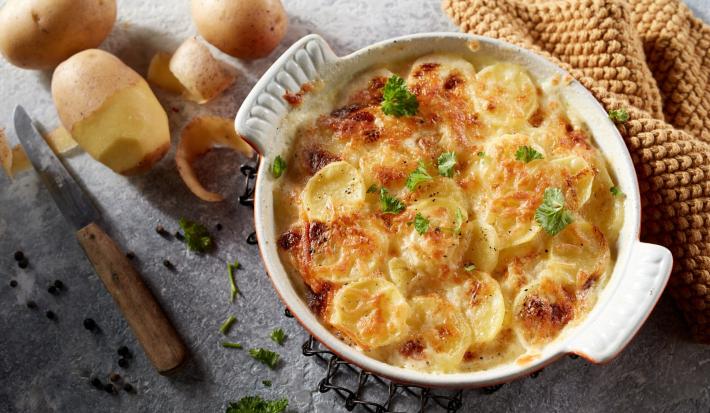 Foto da receita de Batata Gratinada Zero lactose. Observa-se um refratário redondo branco de porcelana com as batatas gratinadas dentro. Ao lado esquerdo, batatas descascadas decoram