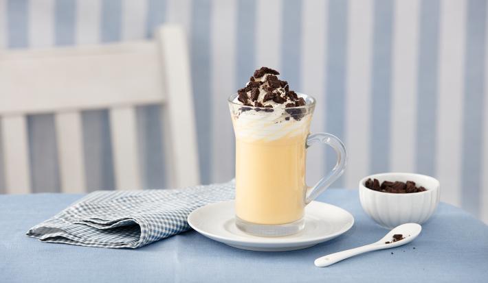 fotografia em tons de azul e branco de uma bancada azul vista de frente, contém um pratinho redondo branco com um copo transparente com a bebida dentro e por cima chantilly e biscoitos triturados para decorar. Ao lado um pano azul e do outro uma colher.