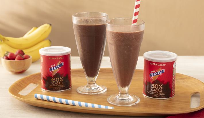 Foto em tons de vermelho da receita de shake saudável feita com Nescau Extra Cacau servida em dois copos altos de vidro sobre uma tábua de madeira com duas latas de nescau e canudos listrados. Ao fundo, bananas e morangos para decorar  sobre uma mesa bege