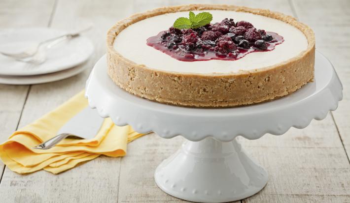 fotografia em tons de branco, bege e roxo de uma bancada cinza vista de frente, contém um suporte para servir sobremesas branco com a torta com calda de frutas vermelhas por cima, ao lado um pano laranja com uma espátula e 2 pratos ao fundo branco.