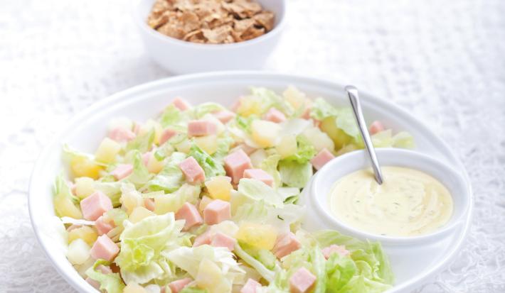 Fotografia em tons de branco e verde de uma bancada de madeira branca com paninho branco, sobre ele um recipiente redondo branco com a salada tropical e com o molho de iogurte. Ao fundo um recipiente redondo branco com o Nesfit Cereal.