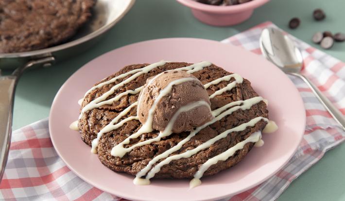 Fotografia em tons de marrom em uma bancada de madeira de cor verde. Ao centro, um pano rosa quadriculado com o cookie dentro. Ao fundo, uma frigideira contendo mais cookie e ao fundo, um pote com gotas de chocolate.