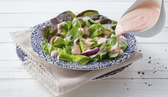 Fotografia em tons de azul, rosa e verde de uma bancada de madeira branca com paninho branco e um prato azul desenhado, sobre ele uma salada de alface com o molho rose e um recipiente despejando o molho sobre a salada.