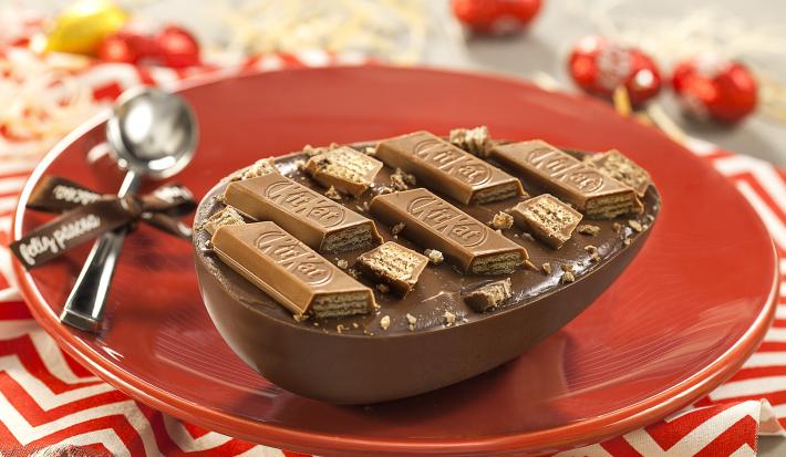 Fotografia em tons de vermelho em uma bancada de madeira cinza, um pano vermelho com listras brancas, um prato vermelho redondo raso com um ovo de Páscoa recheado com ganache e KitKat de Chocolate ao Leite.